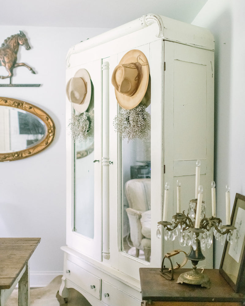 Vintage home decor finds featuring a white antique wardrobe adorned with hats and dried flowers in a charming interior.
