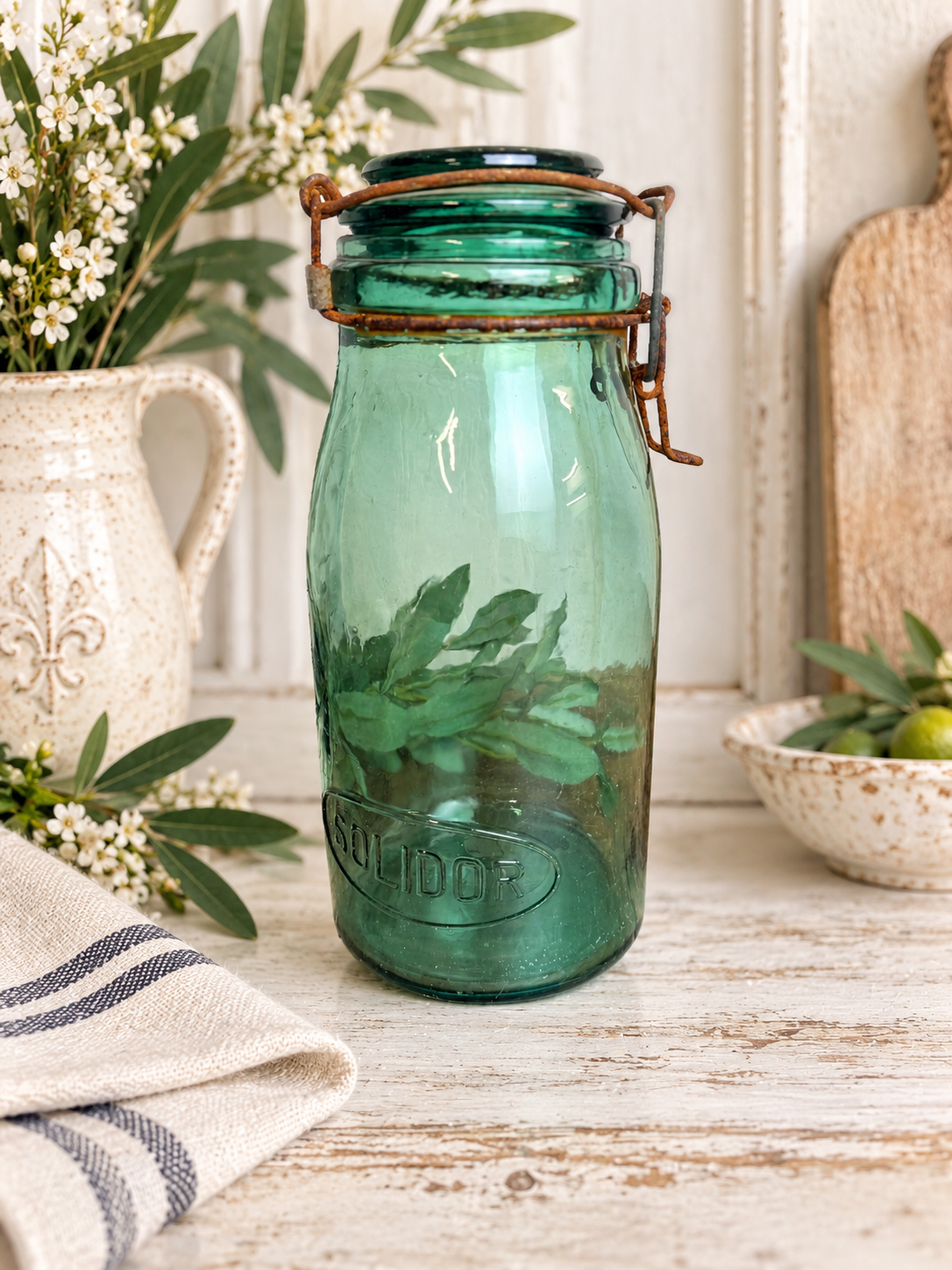 “Green glass jar with a glass lid and wire bail closure on a white wooden table beside a ceramic pitcher.”
