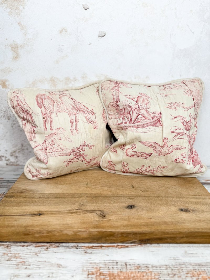 Pair of vintage decorative pillows on a wooden surface, featuring beautiful scenery in red and cream colors.